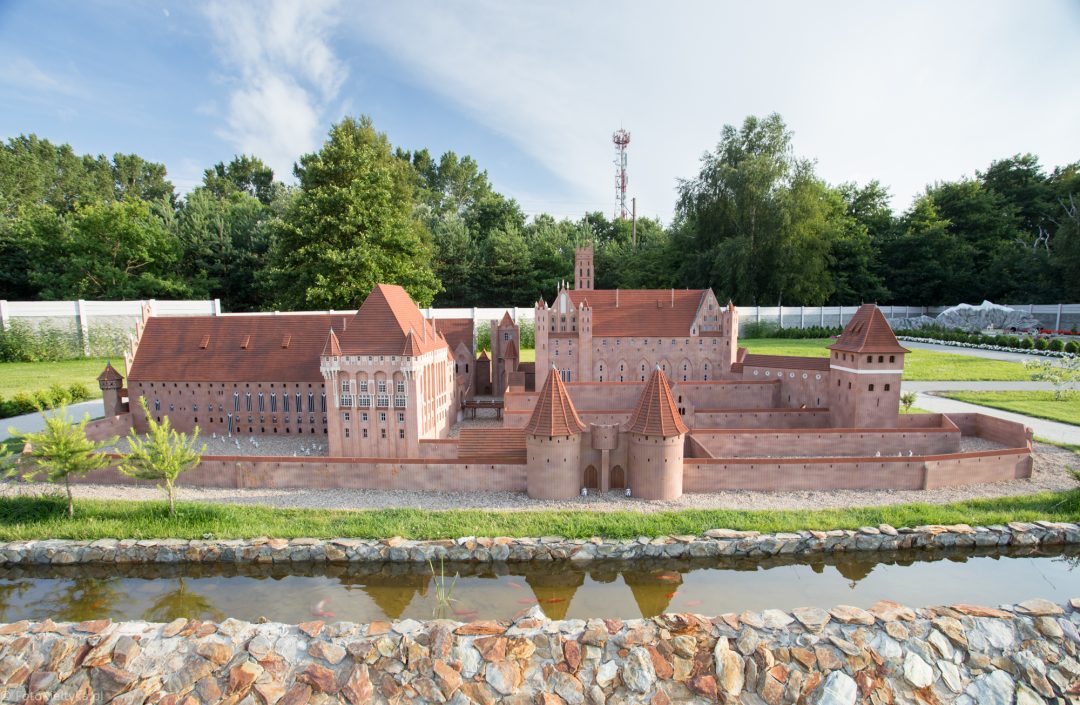 Miniatura rozległej ceglanej budowli z czerwonymi dachami i schodkowymi szczytami.