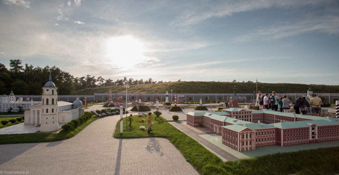Panorama parku miniatur z modelami budowli, alejkami i słońcem nad horyzontem.