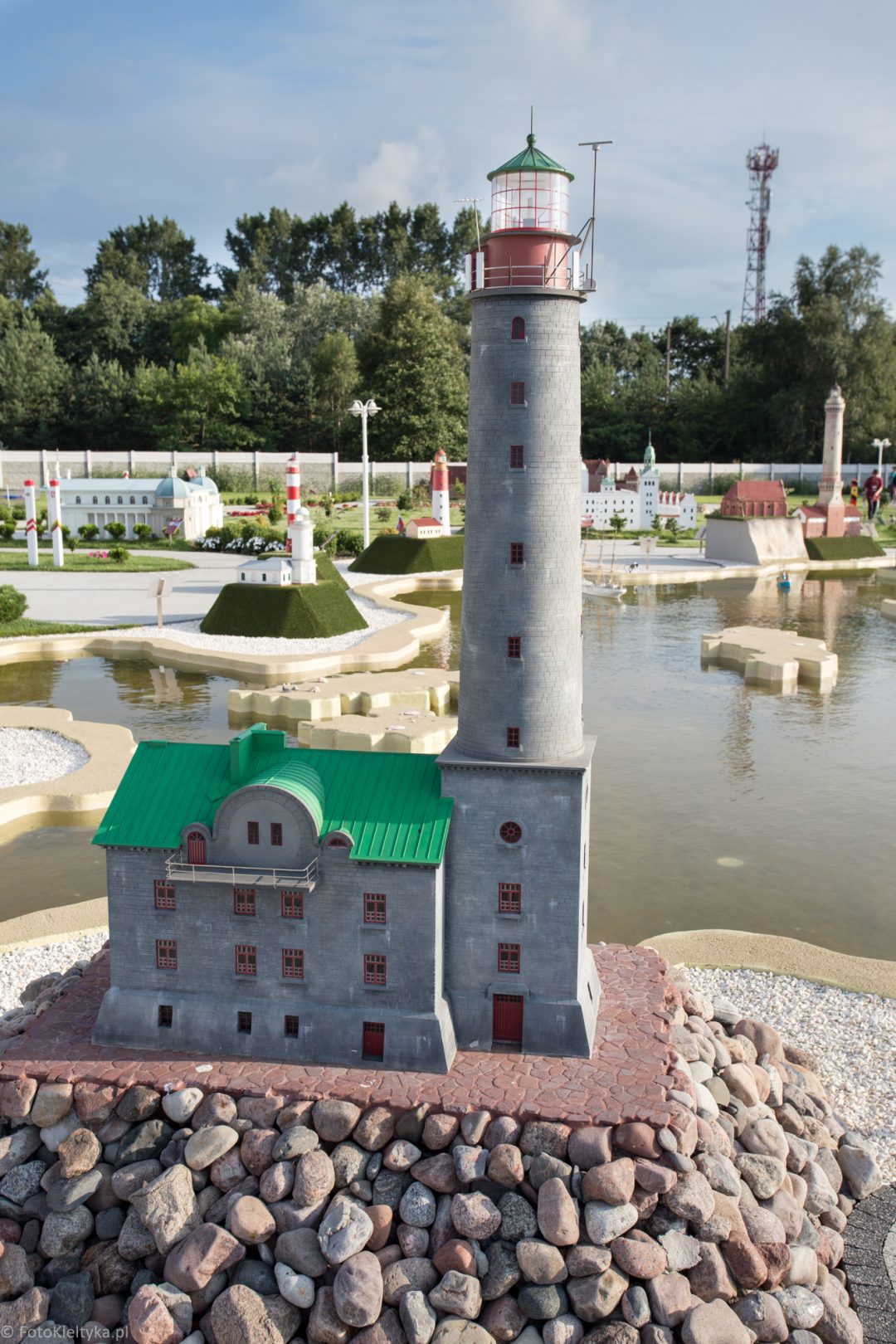 Model jasnej latarni morskiej z czerwonym zwieńczeniem w parku miniatur.