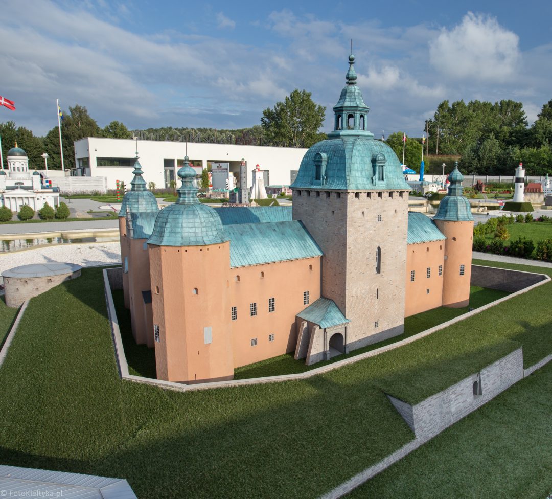 Miniatura ceglanego zamku z narożnymi wieżami i zielonymi dachami, widok z boku.